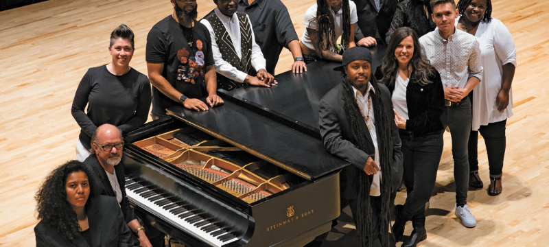 cover photo for magazine with 13 people standing around a grand piano on a concert stage