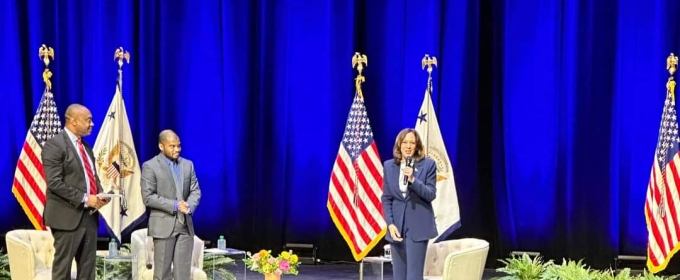 photo of three people on stage with flags, chairs, table